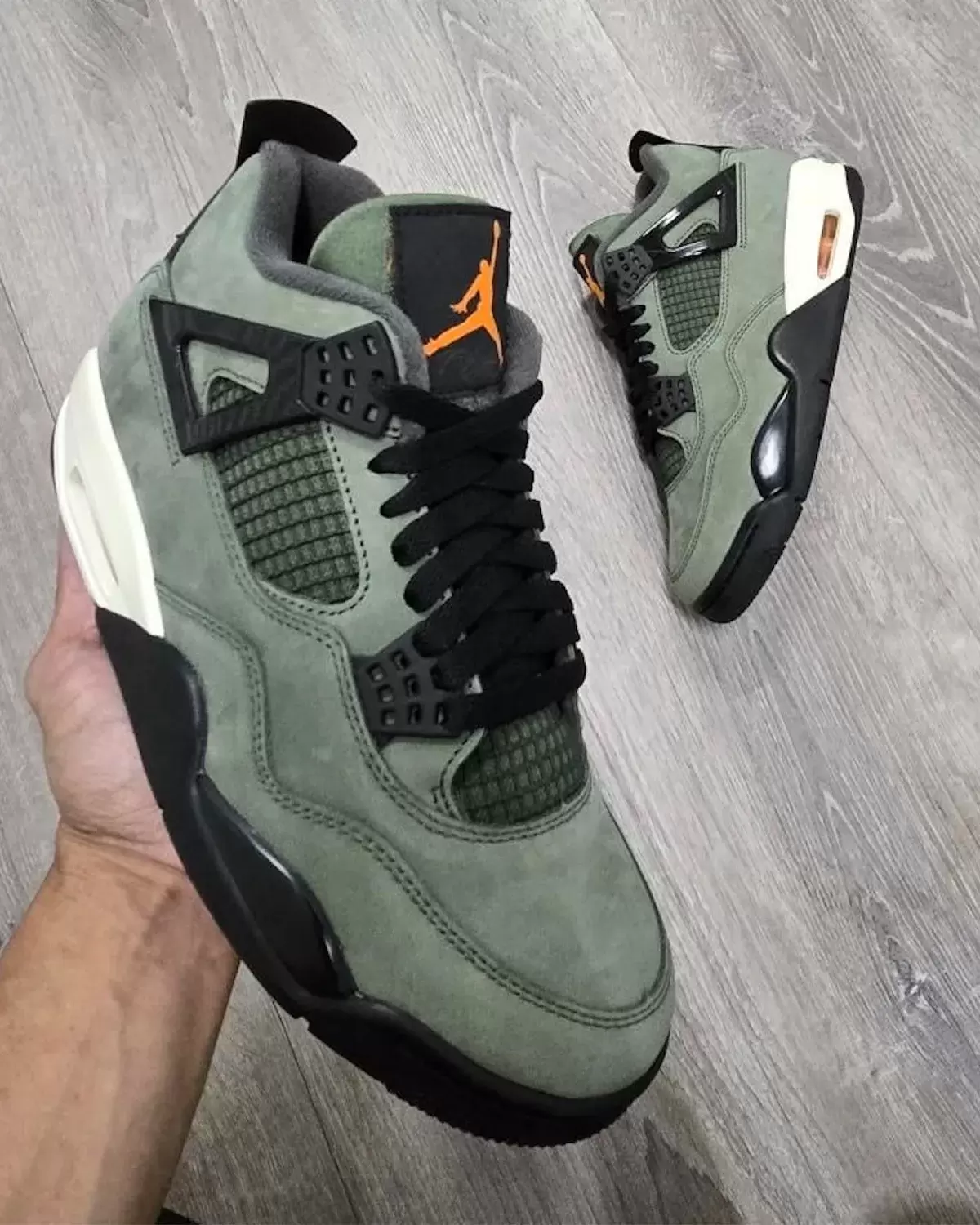 Undefeated Air Jordan 4 2025 sneaker in-hand view showing olive green suede upper and orange Jumpman logo Undefeated Air Jordan 4 2025 sneaker in-hand view showing olive green suede upper and orange Jumpman logo