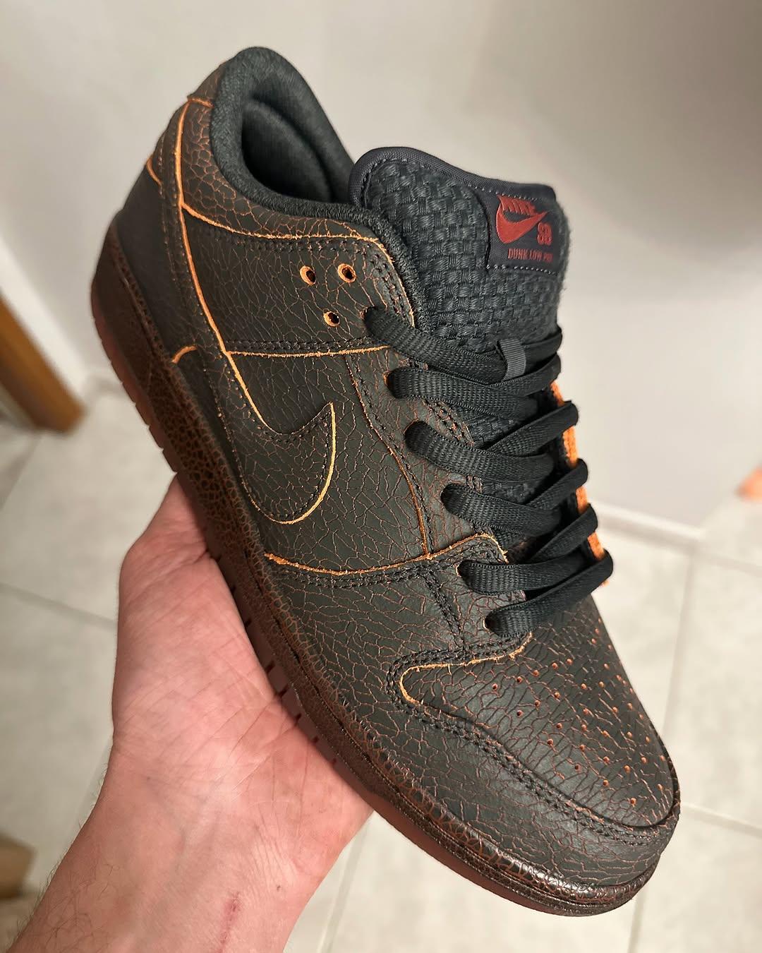 Handheld close-up of the Nike SB Dunk Low Krampus HV1668-001 sneaker. The cracked black leather upper is highlighted by fiery orange stitching, red Nike SB tongue branding, and a dark red sole, showing premium craftsmanship and mythological design inspiration. Handheld close-up of the Nike SB Dunk Low Krampus HV1668-001 sneaker. The cracked black leather upper is highlighted by fiery orange stitching, red Nike SB tongue branding, and a dark red sole, showing premium craftsmanship and mythological design inspiration.