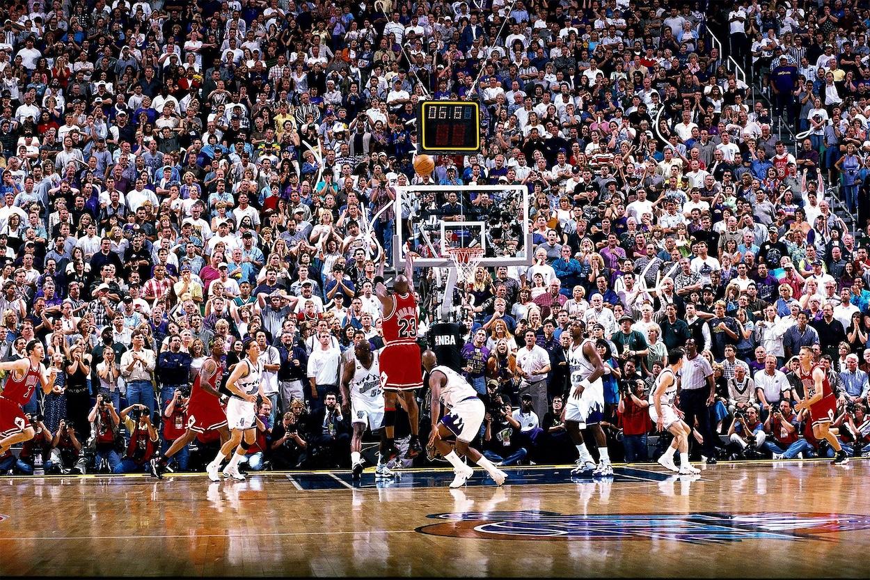 米高-喬丹對猶他爵士隊的最後一投(1998 年) Michael Jordan hits the game-winning shot over Bryon Russell during Game 6 of the 1998 NBA Finals against the Utah Jazz, securing the Chicago Bulls' sixth championship. The legendary moment, known as "The Last Shot," captures Jordan mid-air in his red Bulls jersey, number 23, with the crowd frozen in anticipation.