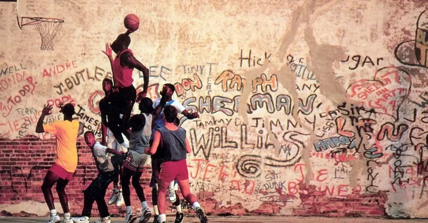 Scene from the 1989 「Michael Jordan』s Playground」 poster, showing a high-flying dunk on an outdoor court with players gathered beneath and a graffiti-covered brick wall in the background.